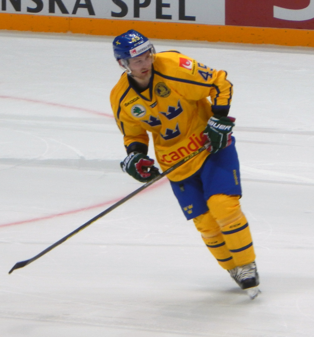 Eishockeyspieler in Bewegung auf dem Eis mit Helm, Handschuhen und Schlittschuhen, der einen Hockeystock hält, mit einem Schild mit Text im Hintergrund.
