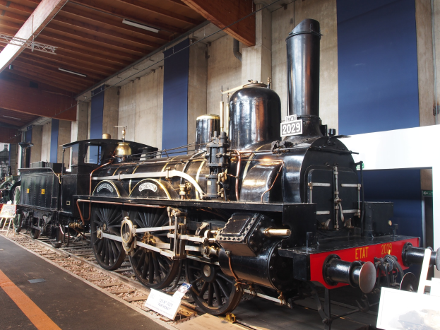 Alte Dampflokomotive in einem Museums Setting mit umliegenden Eisenbahnschienen, Informationsschildern und anderen Ausstellungen, vor einer Wand mit Deckenleuchten.