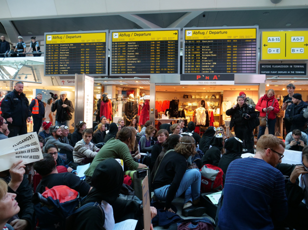 Eine große Gruppe von Menschen in einem Flughafen, einige mit Taschen und Papieren sitzend, andere stehend, mit Texttafeln, Schaufensterpuppen in Kleidern und Deckenleuchten im Hintergrund, was auf eine Protestaktion hindeutet.