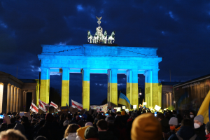 Eine Menschenmenge steht vor dem Brandenburger Tor in Berlin und h├Ąlt Fahnen, Plakate und eine Banner mit Text bei bew├Âlktem Himmel.