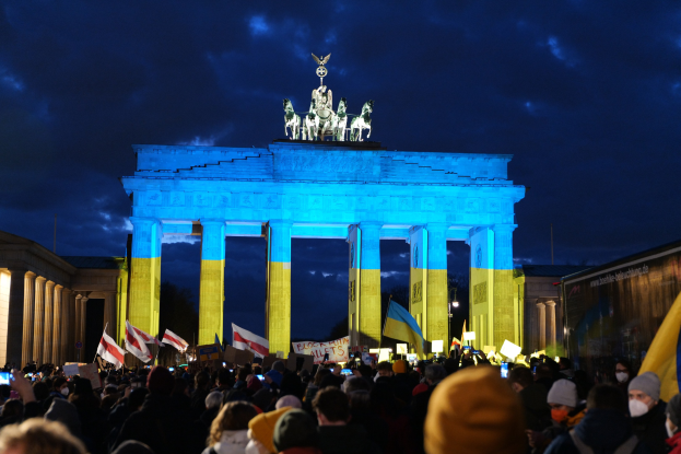 Eine Menschenmenge steht vor dem Brandenburger Tor in Berlin und h├Ąlt Fahnen, Plakate und eine Banner mit Text bei bew├Âlktem Himmel.