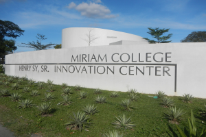 Außenansicht des Miriam College Innovationszentrums mit einer textbedeckten Wand, umgeben von Grün, einem Weg, Bäumen und einem bewölkten Himmel.