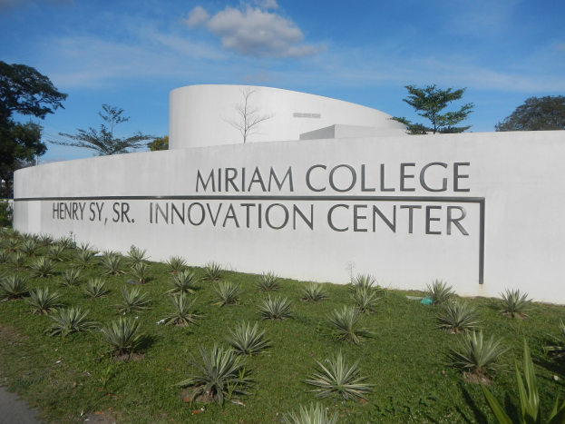 Außenansicht des Miriam College Innovationszentrums mit einer textbedeckten Wand, umgeben von Grün, einem Weg, Bäumen und einem bewölkten Himmel.
