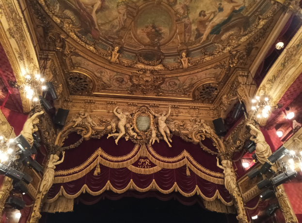 Theaterinnenraum mit roter Gardine, Kronleuchtern, Deckenmalereien, Lichtern und einem Gemälde, das eine Bühne und einige Personen unten zeigt.