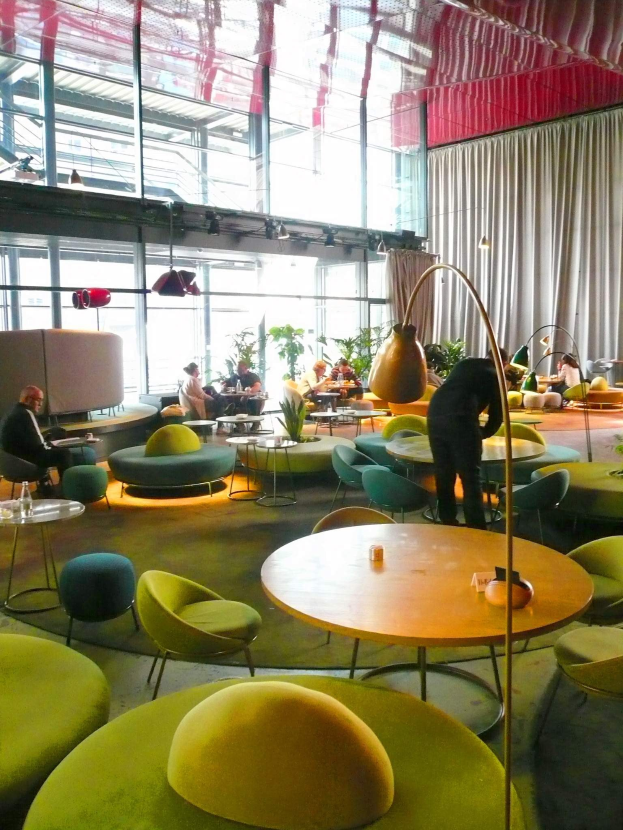 Ein geräumiger Hotel-Salon mit grünen Stühlen, Tischen, Sofas, stehenden Menschen, Pflanzen, einer Stehlampe, Vorhängen, Deckenleuchten und einer gemütlichen Atmosphäre.