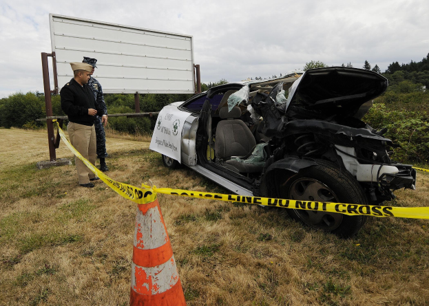 Ein Polizeibeamter in Uniform steht neben einem schwerbeschädigten Auto am Straßenrand, mit eingedellter Motorhaube und zersplitterten Fenstern, unter einem bewölkten Himmel.