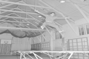 Ein Mann in der Luft, Arme ausgestreckt und Knie gebeugt, auf einem deckenbefestigten Trampolin in einer Turnhalle, dargestellt in Schwarz-Weiß.