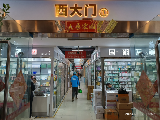 Innenansicht einer chinesischen Apotheke mit Glaswänden, Regalen mit Gegenständen, verstreuten Kartons, einer Pflanze auf der linken Seite und einem Namensschild oben.