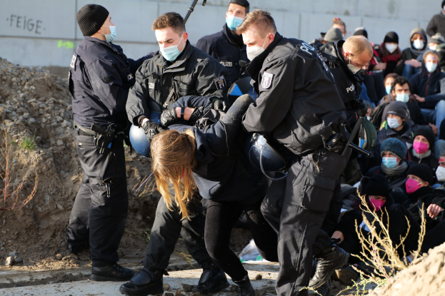 Gruppe maskierter Personen in Mützen, einige bewaffnet, die eine Frau umgeben, die von der Polizei weggeführt wird, mit Pflanzen, Steinen und einer Wand mit Protesttext im Hintergrund.