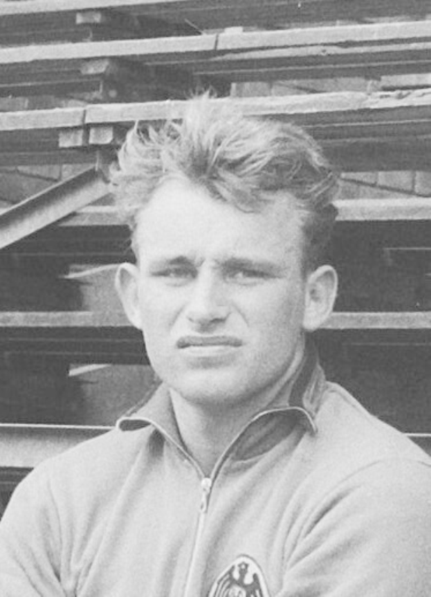 Schwarzes und weißes Foto eines Mannes in einer Jacke, der vor gestapelten Holzplanken steht, der als legendärer deutscher Fußballspieler gilt, der im Alter von 89 Jahren starb.