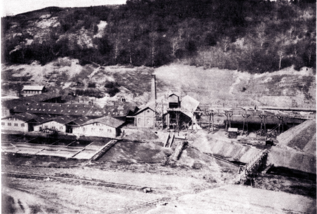 Schwarzes und weißes Foto eines Kohlebergwerks mit Gebäuden, Pfählen und Bäumen im Hintergrund und Text unten.