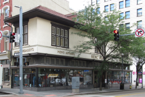 Eine belebte Stadtstraße mit einem geparkten Bus, Gebäuden, Bäumen, Laternen, Verkehrszeichen, Schildern und einem Kaufhaus mit Schaufensterpuppen im Hintergrund unter einem sichtbaren Himmel.