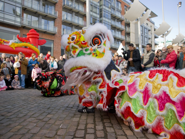 Ein lebendiges chinesisches Neujahrsfest in Amsterdam mit einem Löwen tanzen im Vordergrund und einer Menschenmenge drumherum, einige halten Kameras, vor Buildings, Laternenmasten und einem klaren blauen Himmel.