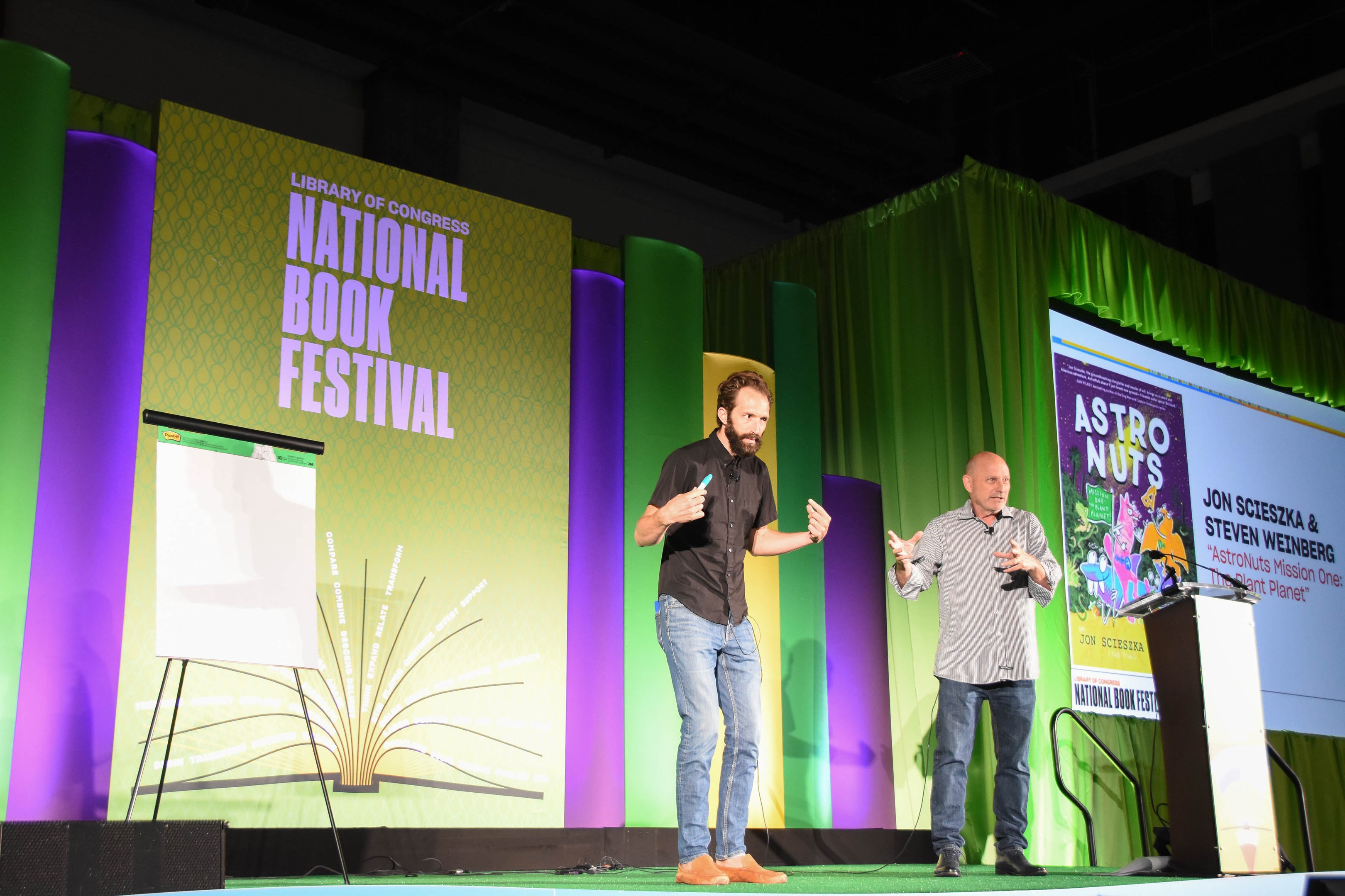 Zwei Männer auf einer Bühne beim Nationalen Buchfestival, mit einem großen Bildschirm dahinter und einem Podium mit Mikrofon auf der rechten Seite.