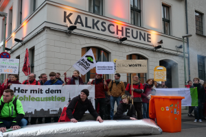 Eine Gruppe von Menschen hält Schilder und Plakate vor einem Gebäude, während zwei Personen im Vordergrund sitzen und ein Müllcontainer rechts davon steht, bei einer Demonstration in Deutschland, mit Gebäuden, Fenstern, Lichtern und Schildern im Hintergrund.