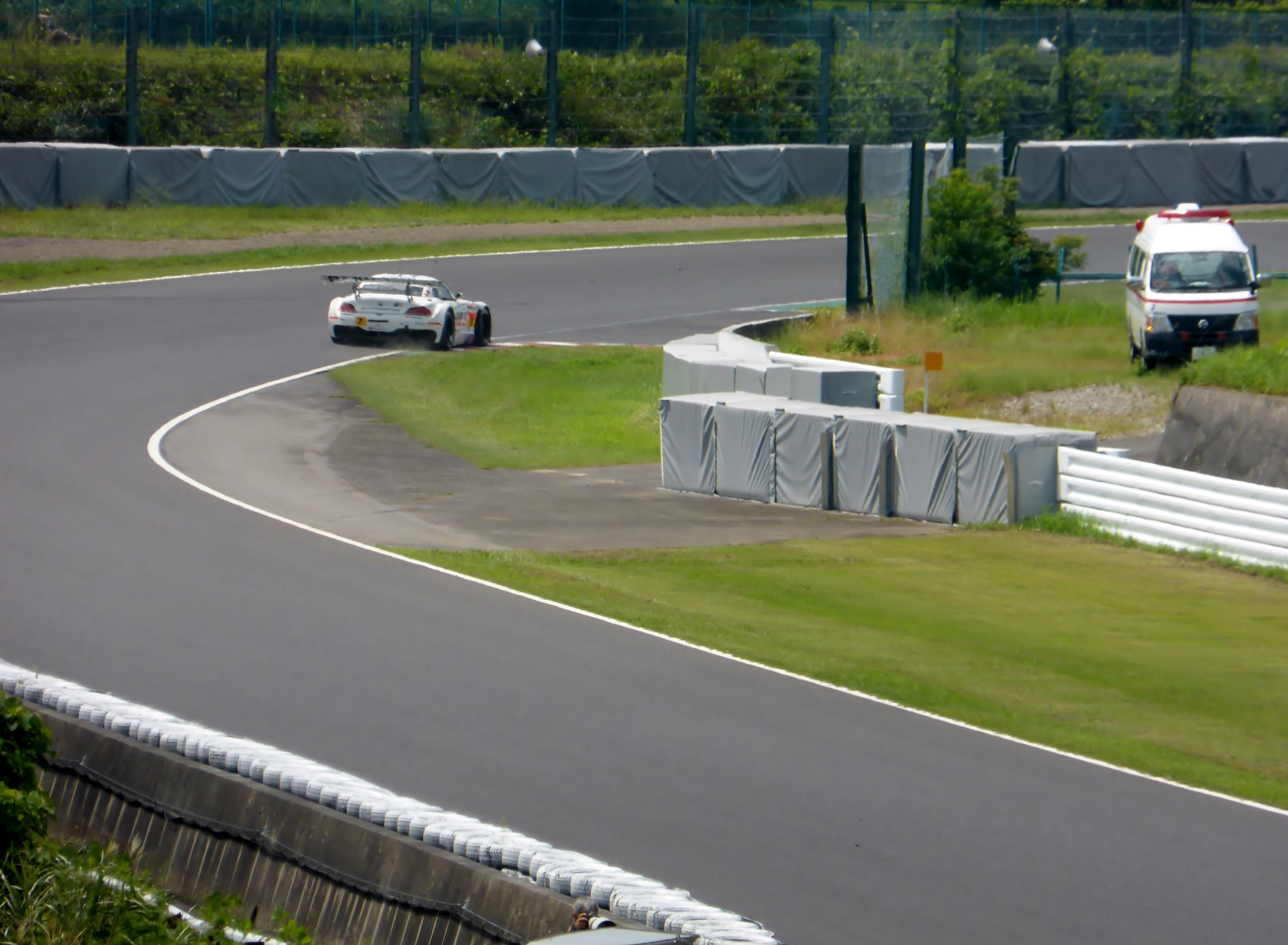Ein Rennwagen fährt auf einer Rennstrecke mit einem Polizeiwagen im Hintergrund, umgeben von Gras, Pflanzen, Bäumen und einer Umzäunung, mit einem Fahrzeug auf der rechten Seite und Text unten.