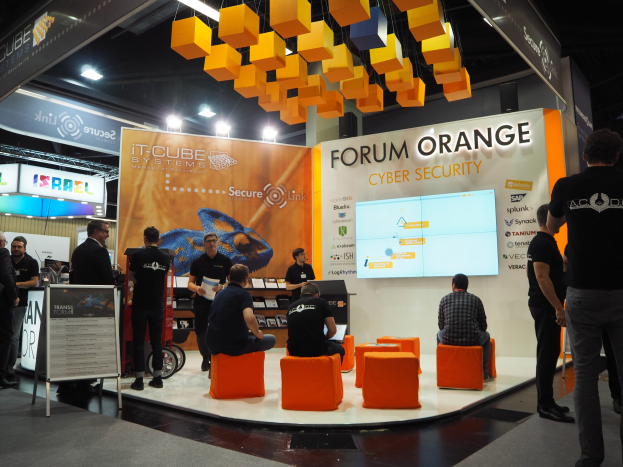 Eine Gruppe von Menschen an einem Messestand für IT-Cube Systems Forum Orange Cyber Security, mit Stühlen, stehenden Besuchern, einer Tafel mit Text, einem Bildschirm, Regalen mit Gegenständen, Schildern, Deckenleuchten und Lampen.