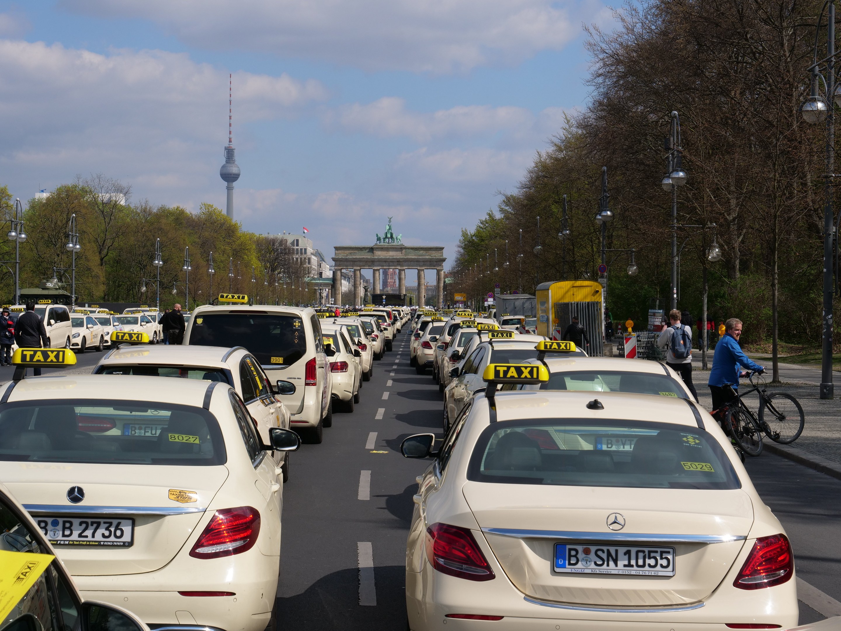 Eine lange Reihe von Taxis, die an einer belebten Straße in Berlin, Deutschland, geparkt sind, mit Fahrzeugen, Radfahrern und Fußgängern, flankiert von Laternenmasten und Bäumen, und Gebäuden, einem Bogen und einem Turm im Hintergrund unter einem bewölkten Himmel.