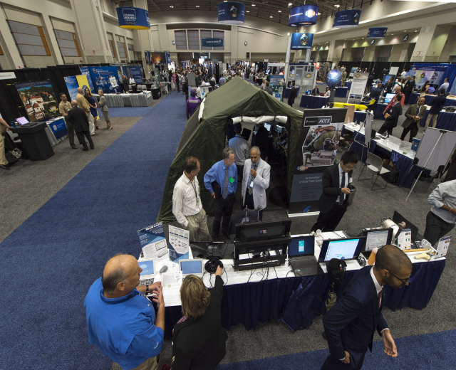 Eine große Gruppe von Menschen auf einer Karriere-Messe steht um einen Tisch mit Laptops, Monitoren und Papieren herum, im Hintergrund sind ein Zelt, Tafeln, Deckenleuchten und Fenster zu sehen.