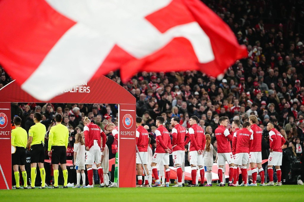 Eine Gruppe von Menschen auf einem Fußballfeld mit einer roten und weißen Flagge im Vordergrund, einem Bogen mit der Aufschrift "Bayern München vs. Bayern München Wetten & Vorschau" im Hintergrund und einer großen Stadionmenge.