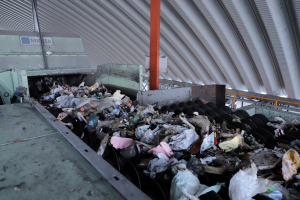 Großer Haufen gemischten Mülls auf einem Förderband in einer Recyclinganlage, mit Maschinen und einem Metalldach im Hintergrund.