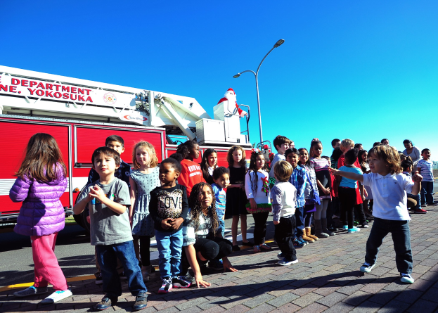 Eine Gruppe von Kindern vor einem Feuerwehrauto, das als Teil der Weihnachtsparade der Yokosuka-Feuerwehr beschriftet ist, mit Laternenmasten, Bäumen und einem klaren blauen Himmel im Hintergrund.