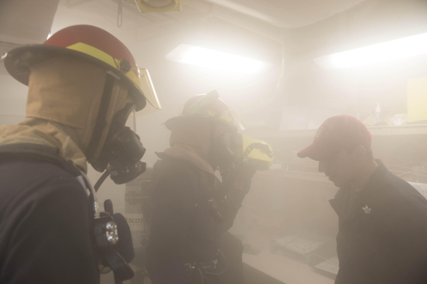 Feuerwehrleute mit Helmen und Rauch aus dem Mund, einer hält eine Kamera, während einer Trainingsrunde mit Hintergrundbeleuchtung und -objekten.