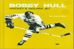 Bobby Hull, ein Eishockeyspieler, hält einen Eishockeyschläger und ein Buch mit dem Titel "Hockey's Golden Jet" von Julian May, trägt einen Handschuh.