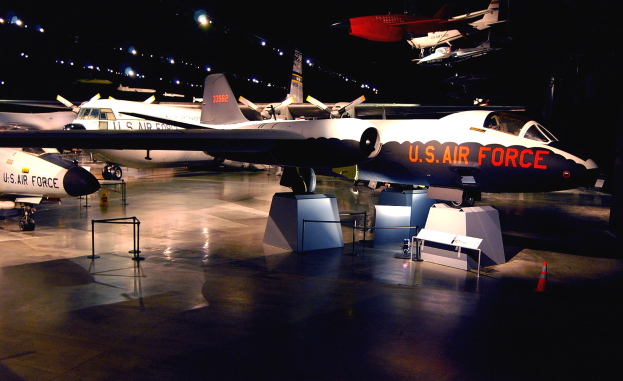 Gruppe von Kampfflugzeugen in einem Museum ausgestellt, beleuchtet von überkopflicht gegen einen dunklen Hintergrund, mit St├Ąnden, Verkehrskegeln und anderen Gegenst├Ąnden auf dem Boden.