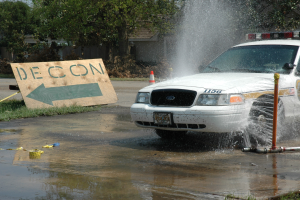 Polizeifahrzeug fährt durch eine Pfütze mit verstreuten Gegenständen, darunter ein Verkehrskegel und eine Tafel mit Text, vor einem Hintergrund aus Bäumen, Gebäuden und einem klaren blauen Himmel.