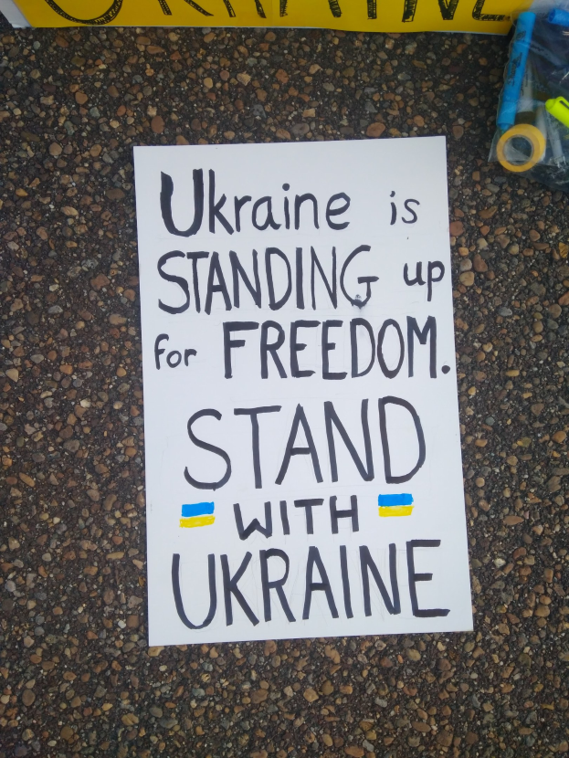 Weißer Zettel mit "Ukraine steht für Freiheit, steht mit Ukraine" Text auf dem Boden vor einer gelben Fahne, mit einer Plastikhülle mit Gegenständen rechts daneben.