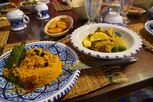Ein Tisch mit Tellern, Wein- und Schüsseln, Krügen, Besteck und Matten, mit Menschen im Hintergrund bei einem traditionellen marokkanischen Essen.
