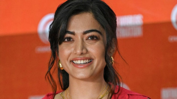 Frau in rotem Kleid mit elegantem Dutt und dezentem Make-up, die in die Kamera lächelt, trägt schwarze High Heels und hat einen hellgelben Banner mit der Aufschrift 'Sonam Kapoors beste Schönheits Looks vom Cannes Film Festival' im Hintergrund.