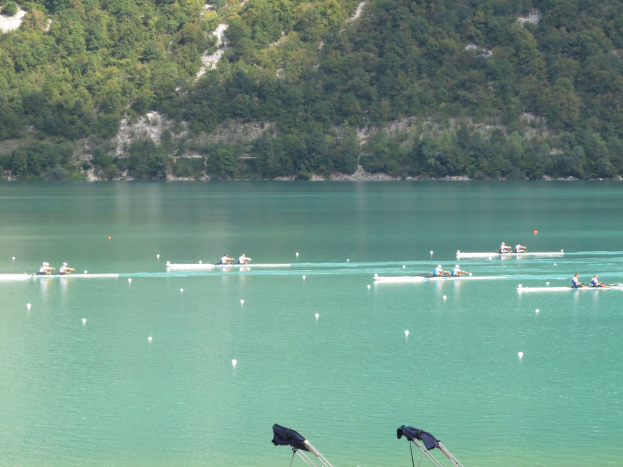 Gruppe von Menschen beim Ruderwettbewerb auf einem Gewässer mit Bäumen und einem Hügel im Hintergrund, Kleider auf Ständen am Boden.