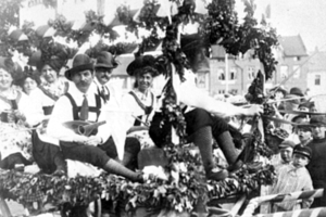 Schwarzes und weißes Bild von Menschen in traditioneller bayrischer Tracht auf einem geschmückten Wagen in einem Umzug, einige spielen Instrumente, mit Gebäuden und einem klaren Himmel im Hintergrund.