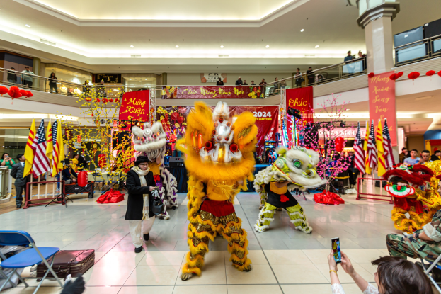 Ein farbenfrohes chinesisches Neujahrsfest in einem Einkaufszentrum mit Menschen in Kostümen, die tanzen, anderen, die auf Stühlen sitzen und Handys halten, sowie Flaggen, Bannern, Dekorationen, Geländern, Säulen, Deckenleuchten und einem Wasserzeichen unten.