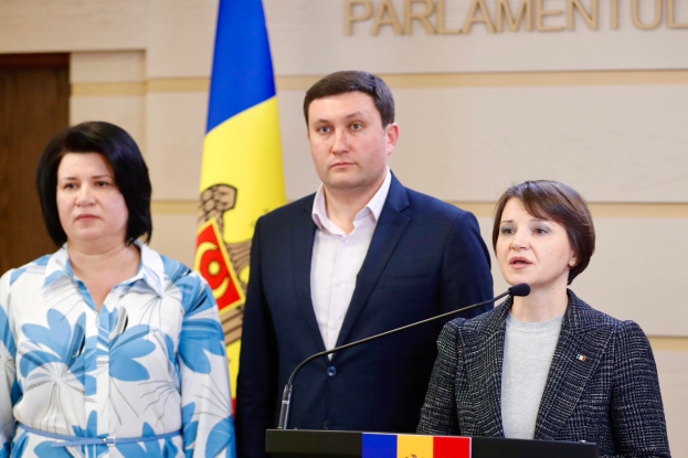 Zwei Frauen und ein Mann stehen an einem Podium vor einer ukrainischen Flagge, wobei der Mann einen schwarzen Blazer und ein weißes Hemd trägt und die Frau rechts ein graues Kleid trägt und über die Ukraine-Krise diskutieren.