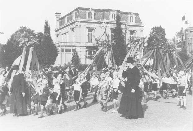 Ein Schwarz-Weiß-Foto von einer Gruppe von Menschen, die eine Straße entlanggehen und Fahnen halten, mit Hüten, Bäumen, Gebäuden und einem Maibaum im Hintergrund.