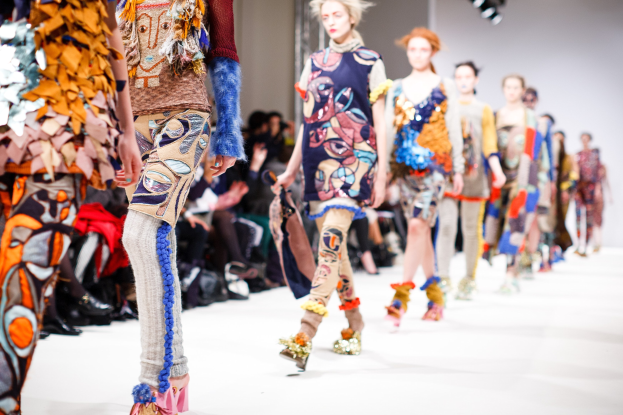 Eine Gruppe Von Frauen Geht Den Laufsteg Bei Der London Fashion Week Herunter, Mit Einer Zuschauerschaft Im Hintergrund Unter Helleren Lichtern.