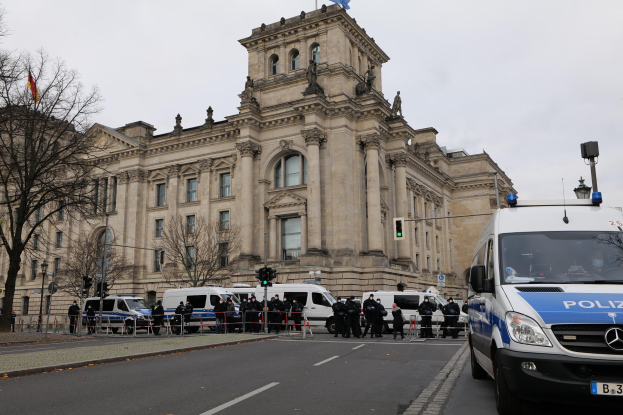 Eine Gruppe von Polizisten steht vor dem Reichstaggebäude in Berlin, Deutschland, mit Fahrzeugen, einem Zaun, Verkehrszeichen, Laternenpfählen, Bäumen und Fahnen im Hintergrund, unter einem klaren Himmel.
