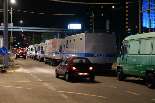 Lange Reihe von Polizeiwagen und -fahrzeugen, die entlang einer Straße bei Nacht mit sichtbarer Straßenverkehr, Straßenschilder, Strommasten, Laternen, Bäume, Gebäude und eine Brücke im Hintergrund unter einem dunklen Himmel geparkt sind.