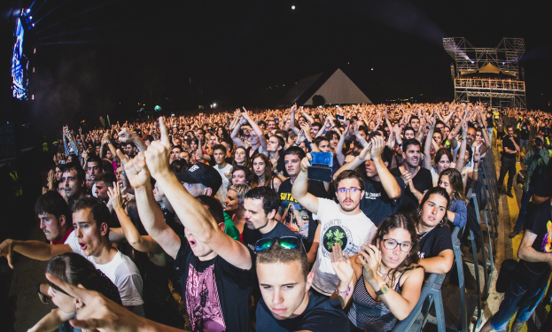 Menschenmenge auf einem Musikfestival mit erhobenen Händen, sitzend und stehend, in der Nähe eines Metallzauns mit Gebäuden und Lichtern im Hintergrund.