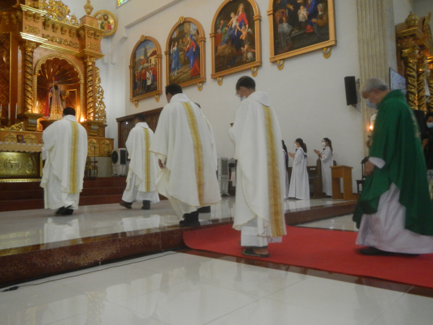 Eine Gruppe von Priestern geht auf einem roten Teppich in einer Kirche, mit Menschen auf dem Boden und auf der rechten Seite, und Fotorahmen an der Wand im Hintergrund.