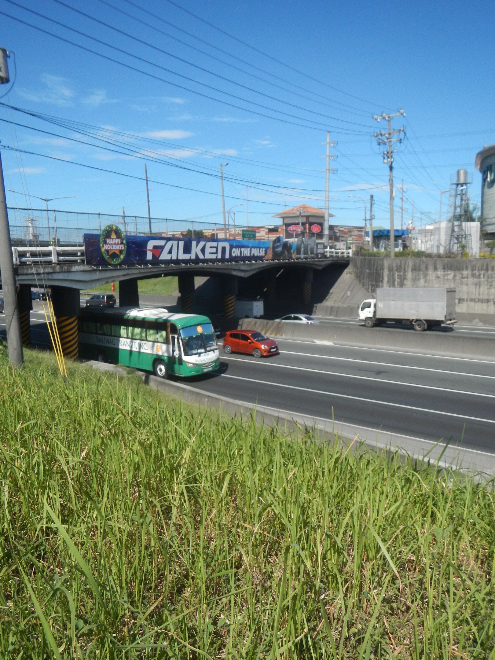 Ein grüner Bus fährt auf einer Autobahn neben hohem Gras, mit einer Brücke, Strommasten, Gebäuden und einem bewölkten Himmel im Hintergrund.