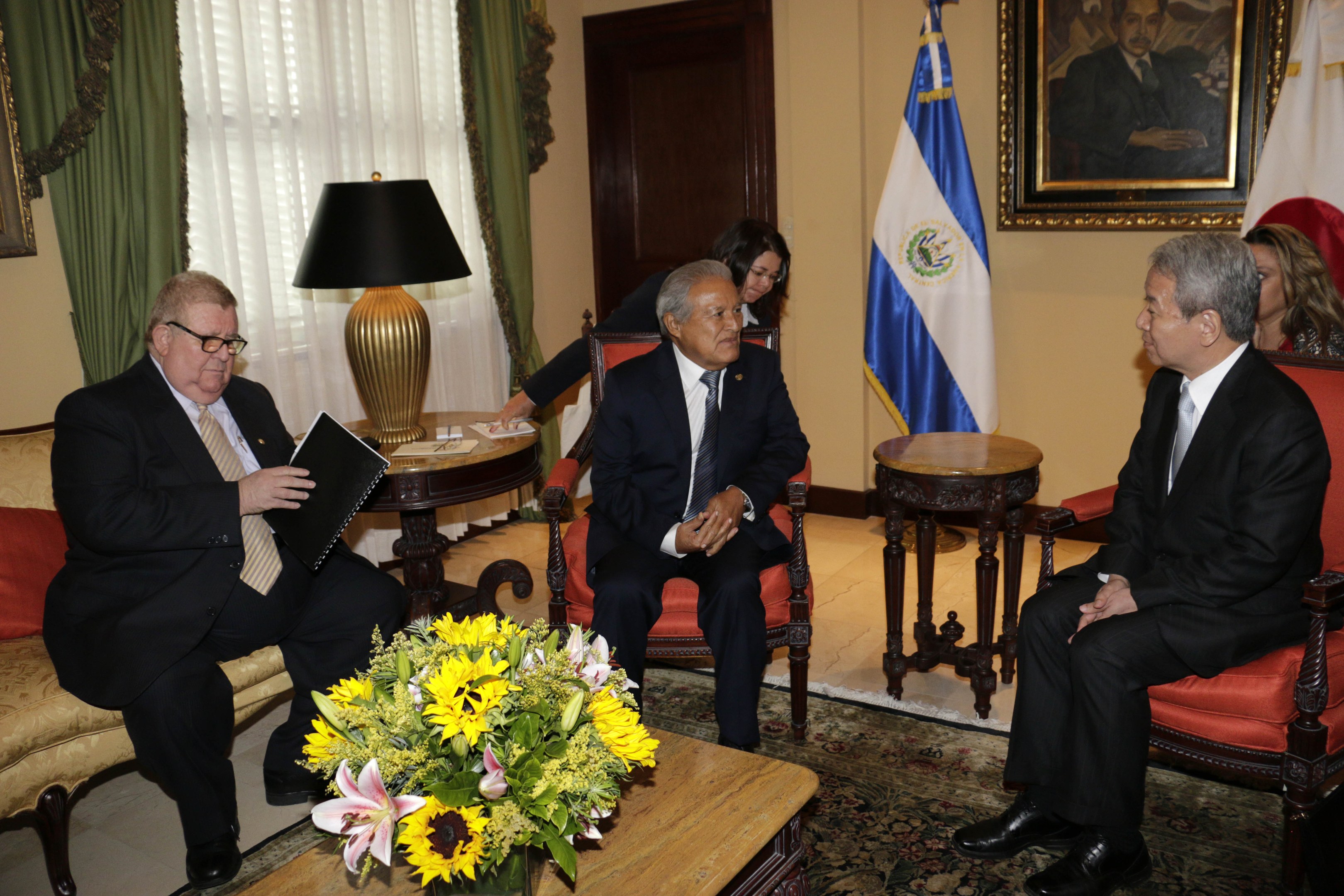 Vier Personen in formeller Kleidung sitzen um einen Tisch mit einer Blumenvase und einer Lampe in einem Raum mit Flaggen, Vorhängen, einer Tür und gerahmten Fotos an der Wand.