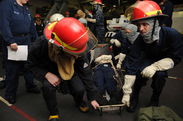 Rettungsteam auf einem Schiffdeck bei der Unterstützung einer Person auf einer Bahre, wobei ein Mitglied Dokumente hält und weiteres Equipment wie Helme, Handschuhe und eine Tasche auf dem Boden zu sehen ist.