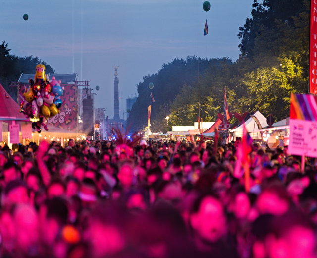 Eine große Menge bei einem nächtlichen Musikfestival mit bunten Lichtern, Ballons, Zelten, Bannern, Fahnen, Bäumen und einem sternenklaren Himmel im Hintergrund.