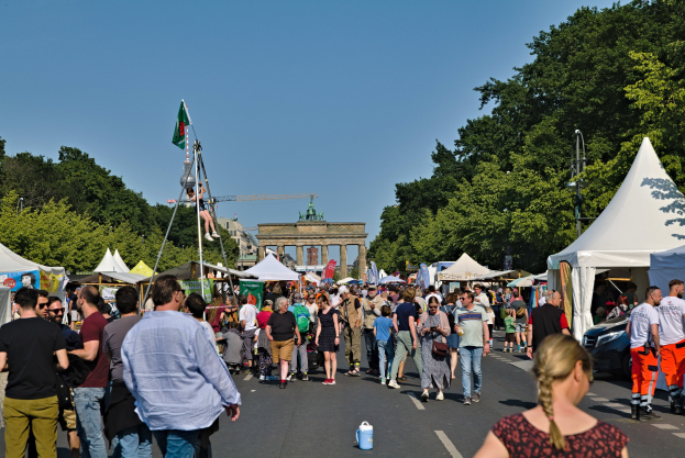 Eine Menge geht eine Straße entlang, gesäumt von Zelten, Fahrzeugen und Bäumen, mit einem Bogen und einem klaren blauen Himmel im Hintergrund und Fahnen an Masten, die das Oktoberfest in München, Deutschland, anzeigen.