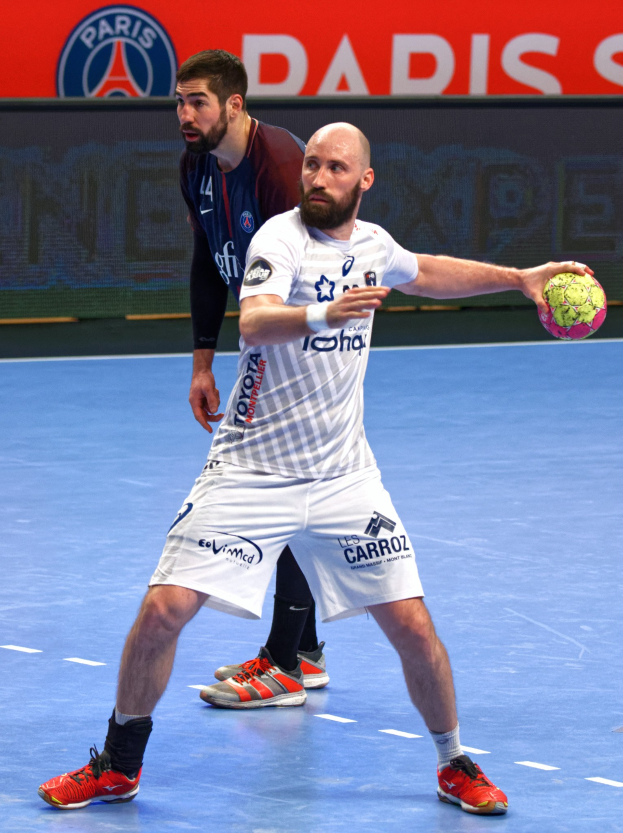 Zwei Männer spielen Handball auf einem Platz, einer hält einen Ball, im Hintergrund ein Schild mit der Aufschrift "Paris Saint-Germain vs Paris Saint-Germain."