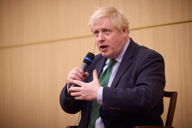 Ein Mann im Anzug und Krawatte sitzt auf einem Stuhl, hält ein Mikrofon und sieht aus, als würde er sich in einer hitzigen Diskussion mit einer Wand im Hintergrund befinden.